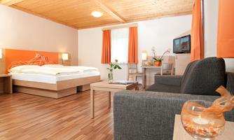 A modern room with a wooden ceiling, a large bed, and a cozy seating area. There are orange curtains and plants that create an inviting atmosphere.