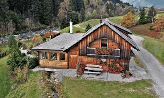 A cozy wooden house surrounded by trees and grass. A small river flows nearby, making the landscape appear idyllic.