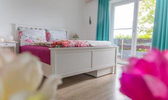 The bedroom is open and bright, the turquoise curtains add a fresh accent. Through the large balcony door, the view goes directly into the greenery, while the flower bed provides splashes of color and coziness. A room where one feels at home immediately.