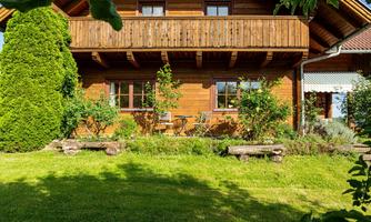 A cozy wooden house with a balcony and a well-maintained garden. Many green plants surround the house, creating a tranquil atmosphere.
