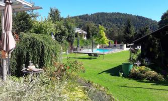 A beautiful garden with well-kept grass and colorful plants. In the background, there is a swimming pool surrounded by trees and mountains.