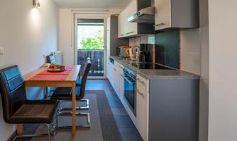 A modern kitchen with a table and chairs. Daylight streams through the window, and the view looks out onto the green surroundings.