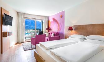 A modern hotel room with a double bed and a cozy reading area. Large windows offer a view of the mountains and plenty of daylight.