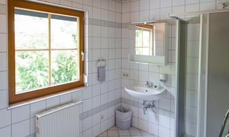 A bright bathroom with white tiles and wooden ceilings. There is a shower, a sink, and a window that lets in plenty of light.