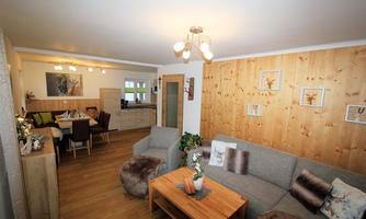 A cozy living room with a gray couch and a wooden table. In the background, a modern kitchen and a dining area can be seen.
