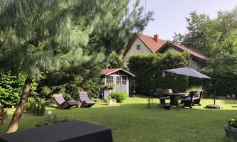 A beautiful garden with green lawn, deck chairs, and a sun umbrella. In the background, a small building and trees can be seen.