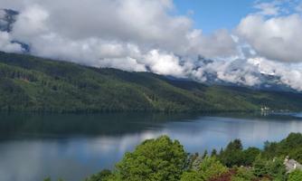 A quiet lake surrounded by green hills and trees. The sky is partly cloudy and is softly reflected in the water.
