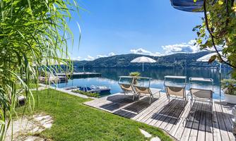 A tranquil lakeside scene with loungers and sun umbrellas on a wooden terrace. In the background, gentle hills and a clear blue sky can be seen.