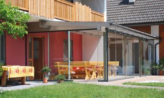 A modern glass veranda with a large table and benches on a lawn. In the background, you can see a cozy house with wooden siding.