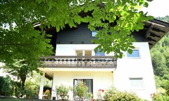 A beautiful house with a balcony and surrounded by plants. The sun is shining and it is a green, quiet environment.
