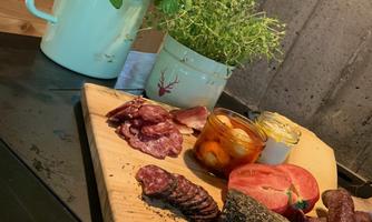 A wooden board with various types of sausage, cheese, and pickled vegetables. Next to it are herb pots in green pots.