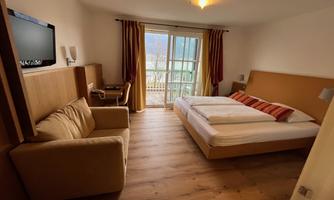 A modern hotel room with a balcony and a large window. There is a double bed, a seating area, and a television.