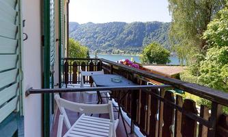 A balcony with a table and a chair that offers a view of a picturesque lake and the surrounding mountains. The scene is surrounded by green trees and conveys a tranquil atmosphere.