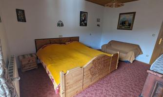 A simple bedroom with a wooden bed and yellow bedding. There is an armchair and wall paintings in the room.