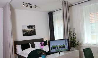 A modern bedroom with a double bed and a television on a white furniture piece. The room is bright with curtains and a small plant arrangement.