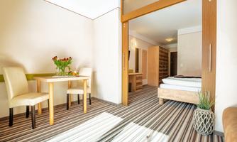 A bright room with two chairs and a table. In the background, there is a bed and a wardrobe.