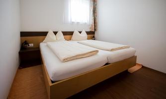 A simple bedroom with two single beds and wooden flooring. The room has a window with daylight and a simple decor.