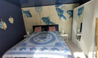 A cozy bedroom with blue wall design and fish motifs. The bed is decorated with a colorful bedspread, and there is a white wardrobe in the room.