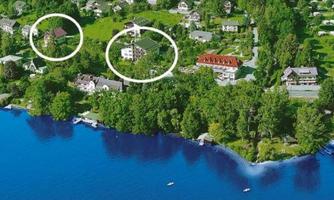 An aerial view of a green, picturesque area with several houses near a clear lake. Boats are visible at the shore, surrounded by lush vegetation.