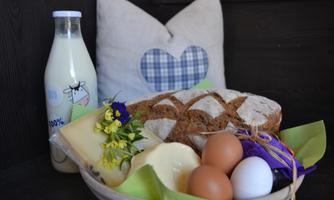 A basket with fresh bread, cheese, eggs, and flowers. Next to it stands a bottle of milk and a pillow in the background.