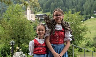 Two girls in traditional costumes are standing in a green landscape. One of the girls is holding a cat on her shoulders.