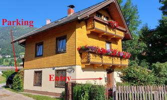 A cozy wooden house with flower boxes and a balcony. The parking area and the entrance are clearly marked.