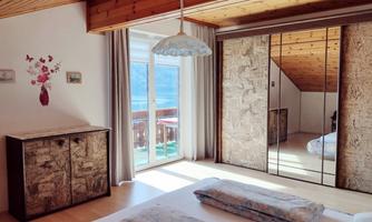 A bright bedroom with a wooden ceiling and large windows. There is a bed with pillows and a wardrobe with a mirror.