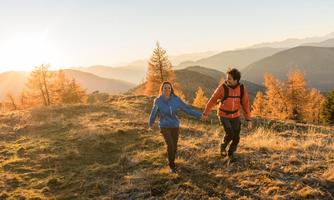 A couple is hiking in the mountains during a sunset. The landscape is surrounded by autumn trees and gentle hills.