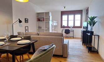 A modern living room with light decor and wooden flooring. There is a dining area, a comfortable sofa, and large windows that let in plenty of light.