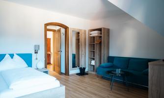 A modern room with a comfortable bed and a blue couch. The space is brightly and friendly furnished, with wooden flooring and a wardrobe.