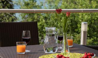 A beautifully set table on a terrace overlooking the water. There are drinks, fresh fruit, and flowers in a vase.