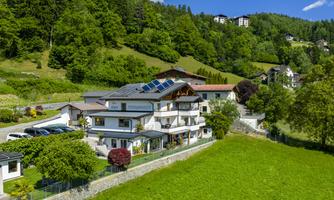 A modern house with solar panels, surrounded by lush greenery and gentle hills. The clear blue sky creates a peaceful atmosphere.