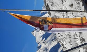 A flag flutters in the wind in front of a snow-covered mountain landscape. In the background, clear blue skies and distinctive peaks are visible.