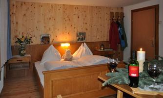 A cozy bedroom with wooden paneling and a double bed. On the table, there are wine and a candle, while a plush toy sits on the bed.