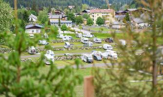 A campsite with various motorhomes in a green environment. In the background, some buildings and trees are visible.