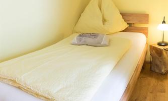 A simple bedroom with a single bed and yellow walls. There is a lamp on the bedside table, and a picture hangs in the background.