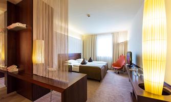 A modern hotel room with two beds, a stylish table, and a cozy armchair. Large windows let in plenty of daylight.