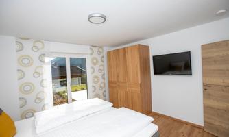 A modern bedroom with a double bed and a large window. A TV is mounted on the wall and the floor is made of wood.