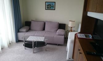 A cozy living space with a bright sofa and a glass table. In the background, there is a television and a window with curtains.