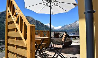 A cozy terrace with a table and chairs, covered by a sun umbrella. In the background, majestic mountains and a blue sky can be seen.