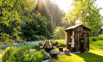 A cozy wooden shed stands by the shore of a calm river, surrounded by lush greenery. The sun shines through the trees, creating an inviting atmosphere.