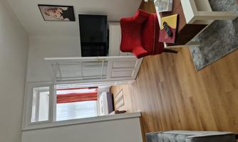 A cozy living room with a red armchair, a television, and a small table. Large windows provide plenty of daylight and a modern decor.