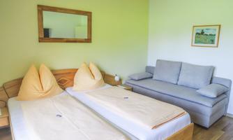 A simple room with two single beds and a gray sofa. The walls are painted in a soft green shade, and a mirror complements the decor.