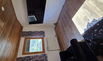 A cozy room with a black armchair and a large television on the wall. The windows provide daylight and the floor is made of wood.