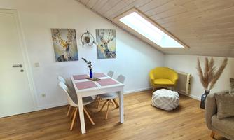 A bright room with a dining table and white chairs. A yellow seating area and decorative pictures on the wall complete the cozy atmosphere.