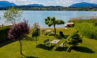 A tranquil lakeside landscape with a clear lake and green meadows. In the foreground are trees and tables that invite relaxation.
