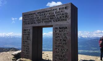 A monumental gate made of metal stands on a mountain peak. In the background, snow-covered mountains and a blue sky are visible.