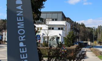A sign with the inscription "Seepromenade" is in the foreground. In the background, modern buildings and a green landscape can be seen.