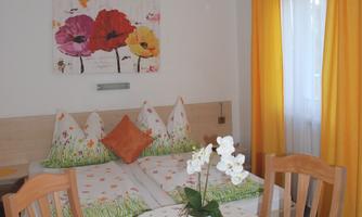 A cozy room with a table, chairs, and colorful cushions. A colorful painting of flowers hangs on the wall.