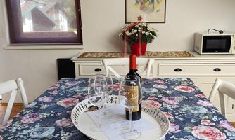 A cozy dining table with a floral tablecloth, on which a bottle of wine and glasses are placed. In the background, plants and a small refrigerator can be seen.